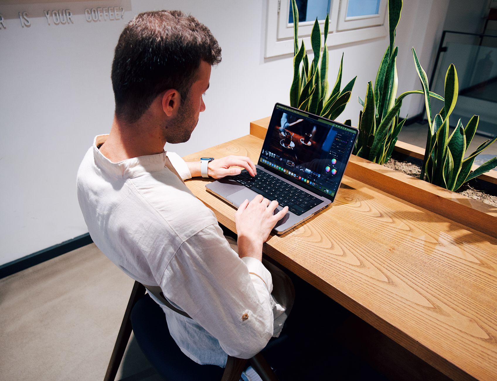 Un homme en chemise claire travaille sur un ordinateur portable posé sur un bureau en bois, à côté de grandes plantes vertes.